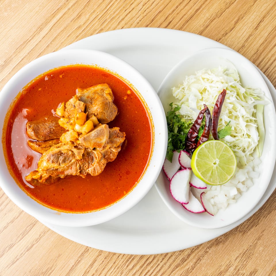 Red chile stew with tender meat served alongside shredded cabbage, radishes, lime, cilantro, and dried chilies.