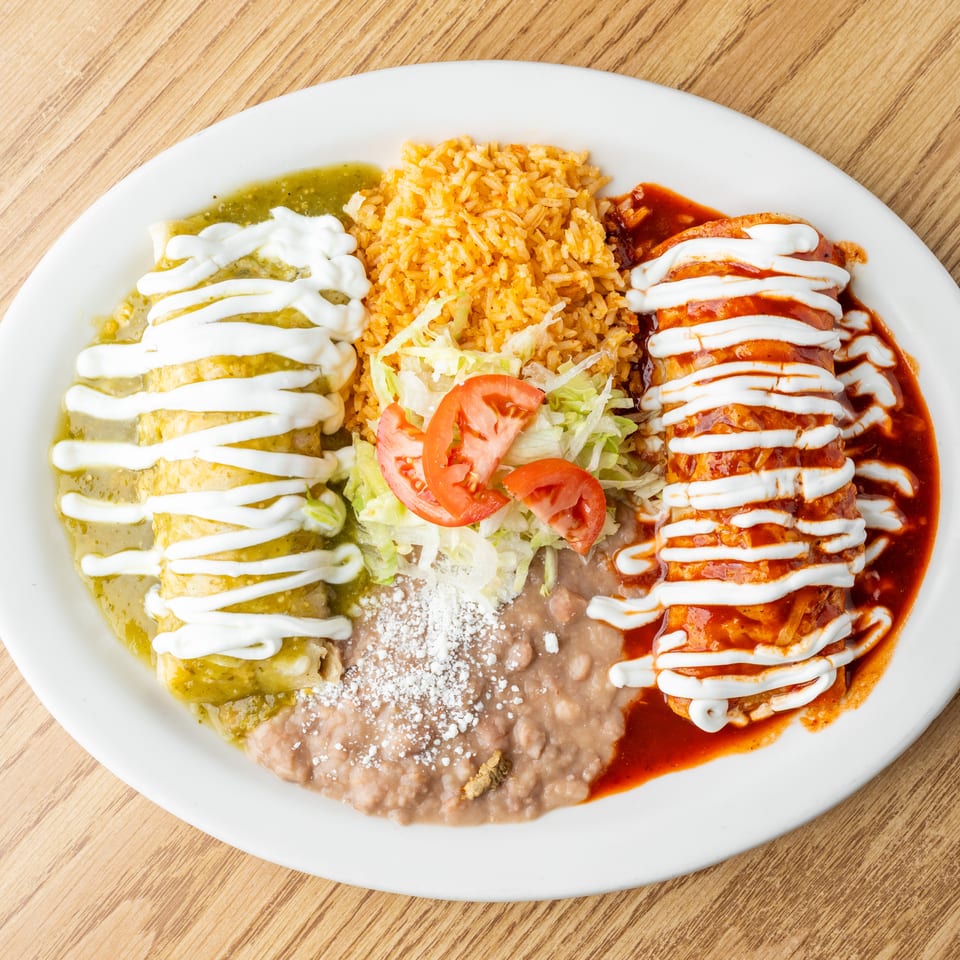 Mexican platter with enchiladas in green and red sauce, rice, refried beans, lettuce, tomatoes, and crema.