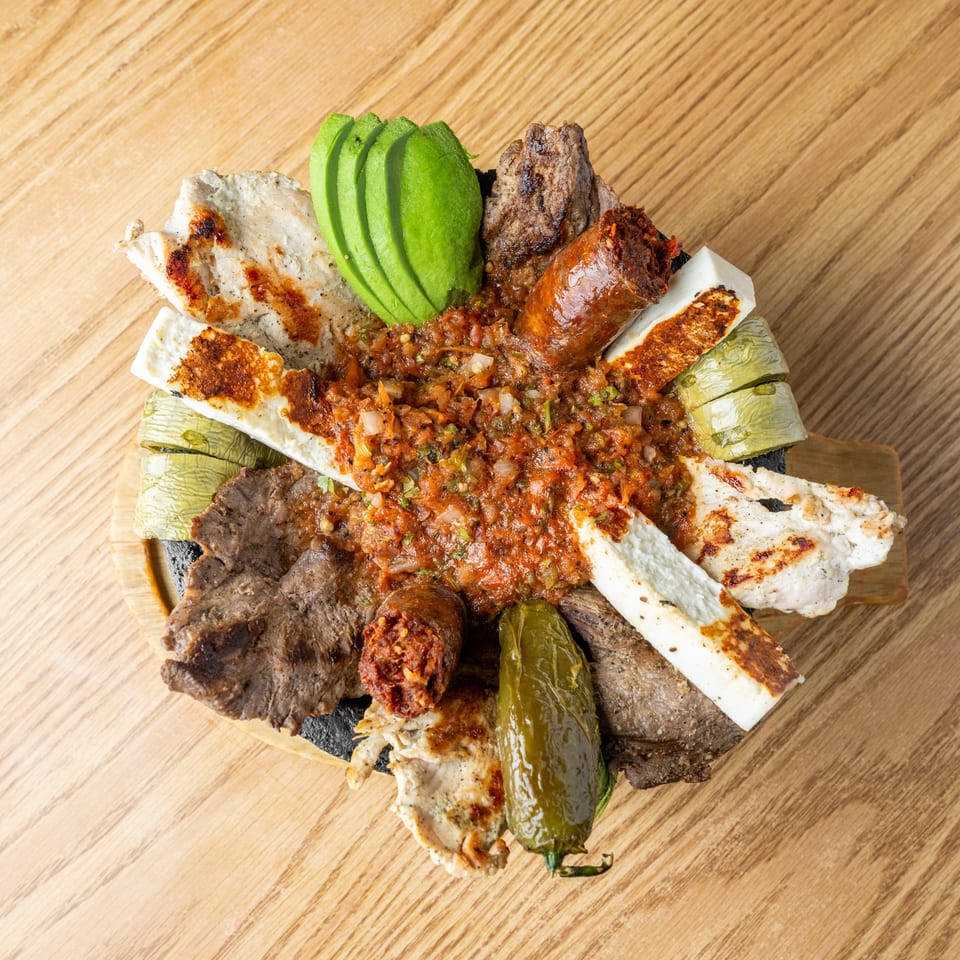 Grilled meat platter with chicken, beef, sausage, halloumi, avocado, grilled vegetables, and spicy tomato relish on a wooden table.