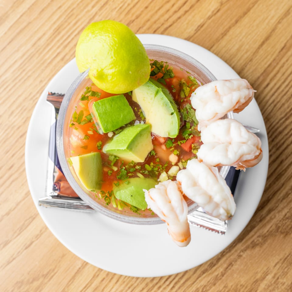 Shrimp cocktail topped with avocado, cilantro, tomato, and lime, served in a glass bowl on a white plate.